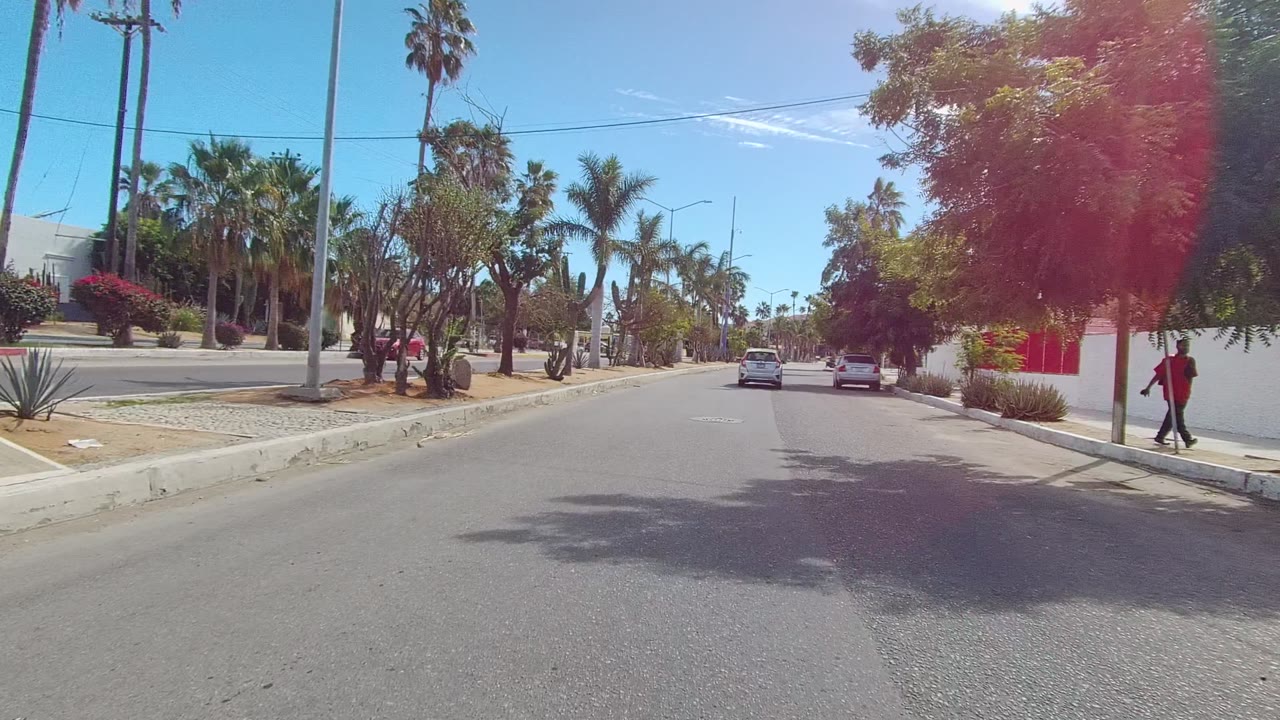 East Cape Los Cabos street frame with a parked silver car near the right curb, palms and cacti along white walls, red trim, and a distant person in red walking on the sidewalk.
