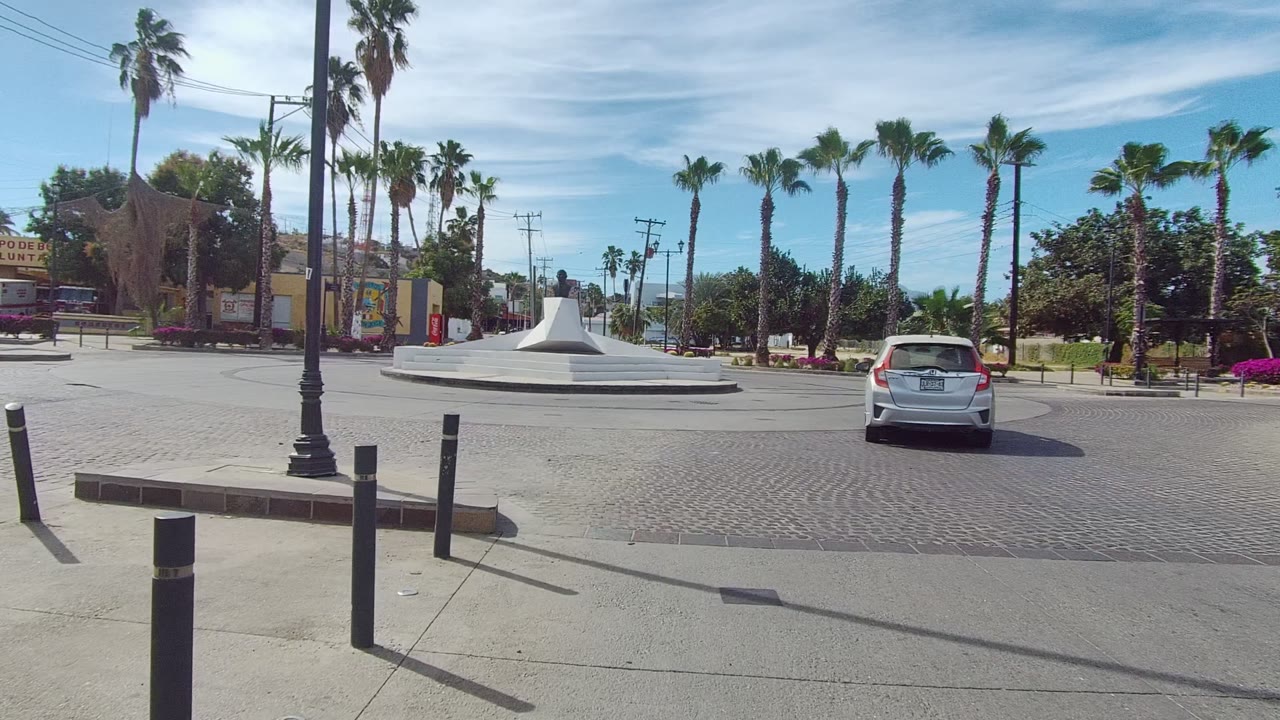 Wide Los Cabos street frame with a white car approaching a low white circular traffic island, tall palms, cobblestone paving, buildings, and no visible people.