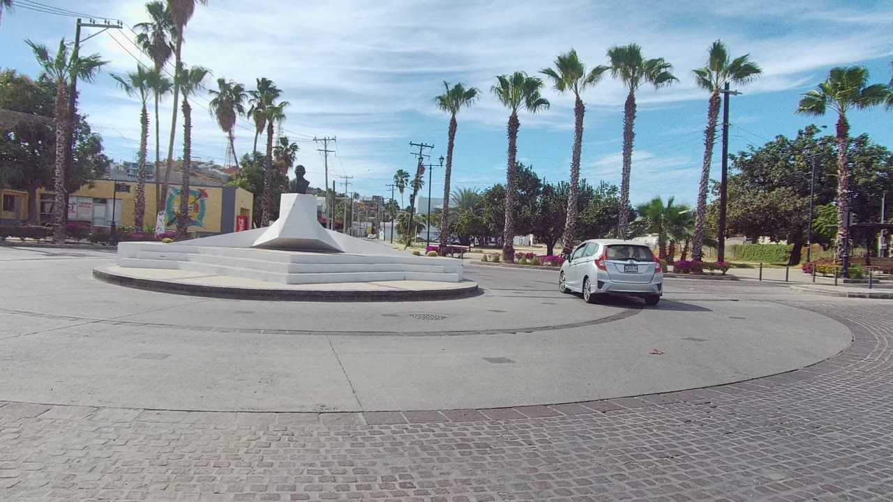 Wide Los Cabos roundabout frame with the white car near the right edge, a sculptural white traffic island, palm trees, buildings, and no visible people.