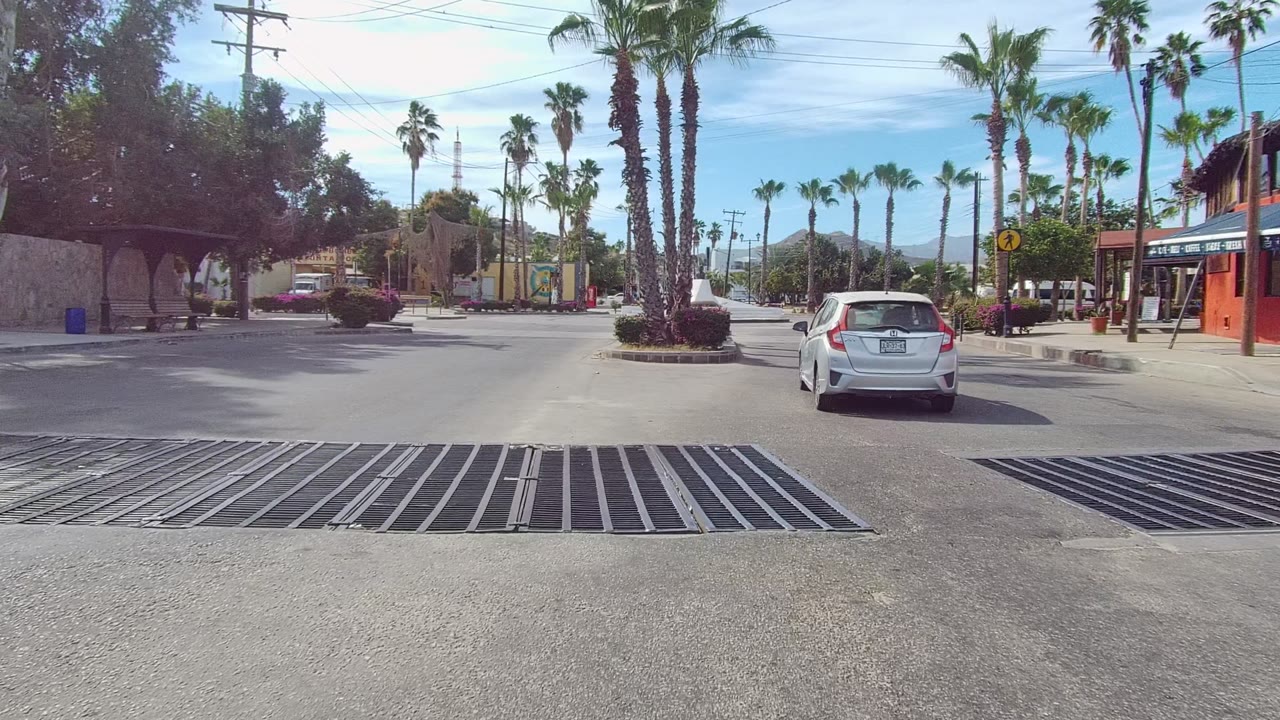 A white hatchback approaches a roundabout in Los Cabos, Baja California Sur, with drainage grates in the foreground, palms and buildings ahead, and no visible people.