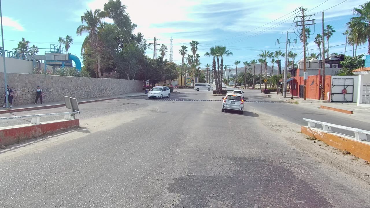 Los Cabos road scene approaching an urban intersection, with a white car ahead, another car at left, palm trees, utility poles, sidewalks, and red-orange building fronts.