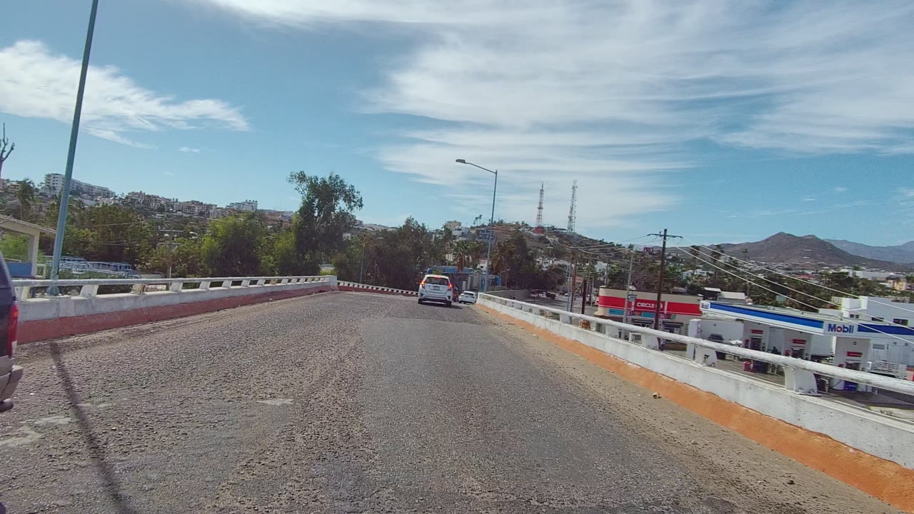 A road-level view in East Cape Los Cabos shows a curving asphalt bridge with concrete barriers, cars ahead, hills in the distance, and no visible people.