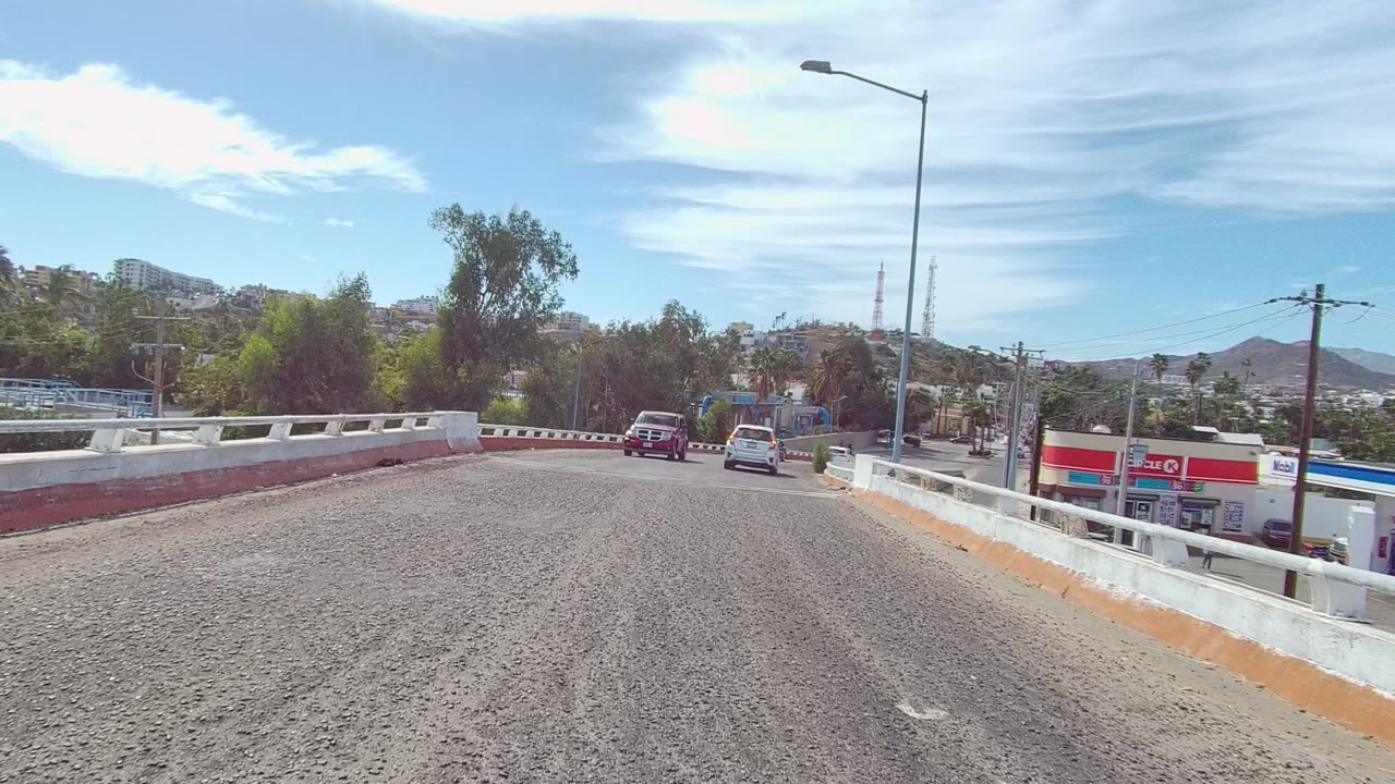 A later bridge view in East Cape Los Cabos shows vehicles spaced along the curve, a red roadside gas station sign and canopy to the right, hills beyond, and no visible people.