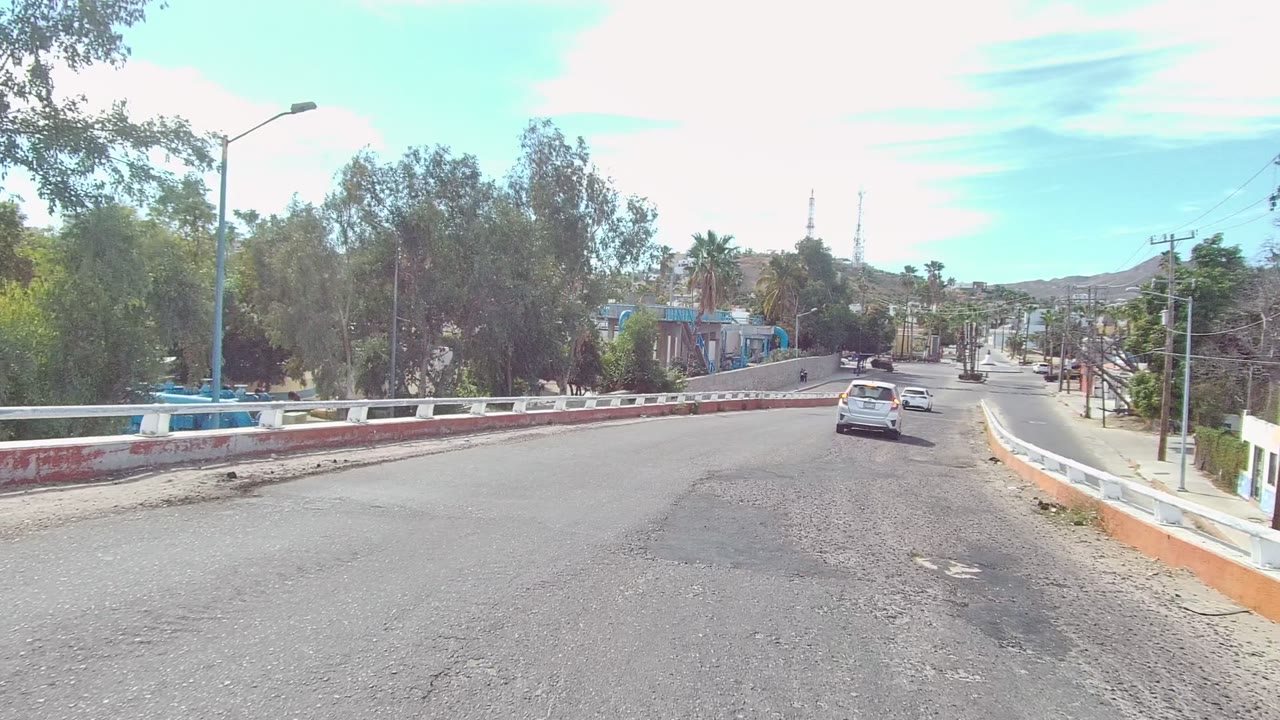Opening view in East Cape Los Cabos, Baja California Sur, with a white car ahead on a curving paved road, red and white barriers, trees, streetlights, and no visible people.