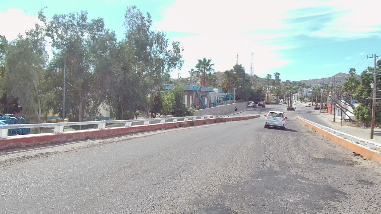 Paved road bending right with a white car, roadside barriers, trees, and buildings.