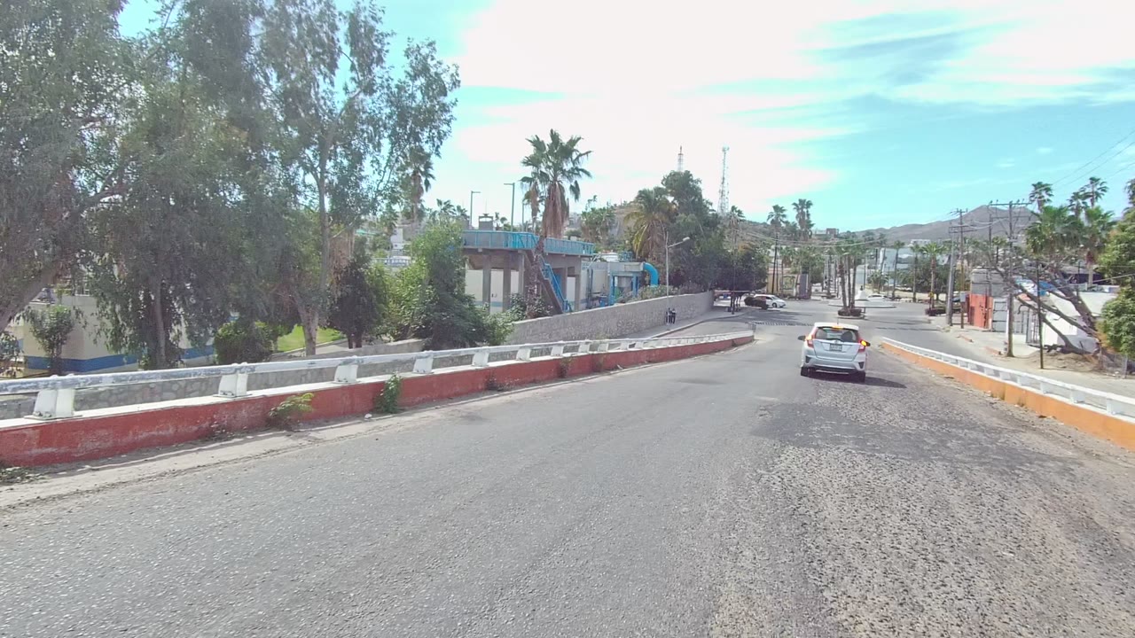 Mid-clip view in East Cape Los Cabos, Baja California Sur, with a white car centered ahead, a red-and-white barrier at left, orange roadside barrier at right, palms, poles, buildings, and no visible people.