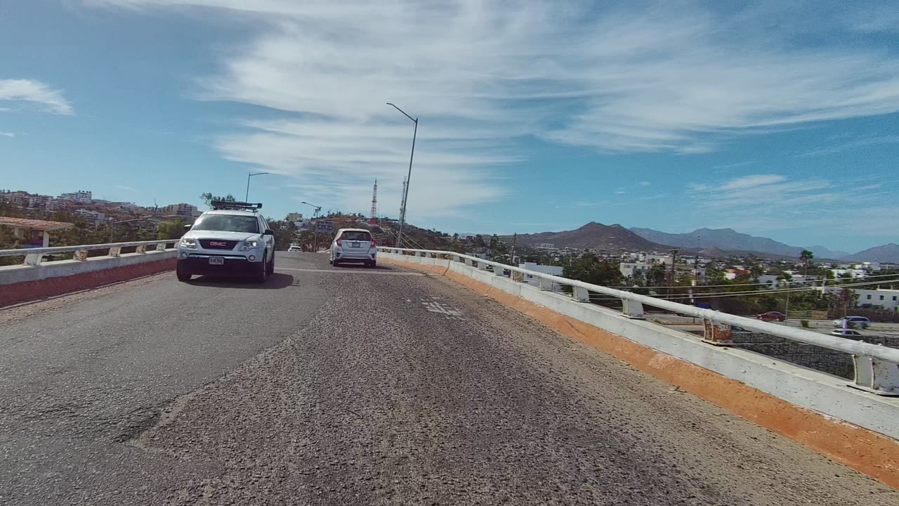 Opening frame of vehicles driving on a bridge road in Los Cabos, Baja California Sur, with concrete guardrails, hillside buildings, mountains, high clouds, and no visible people.