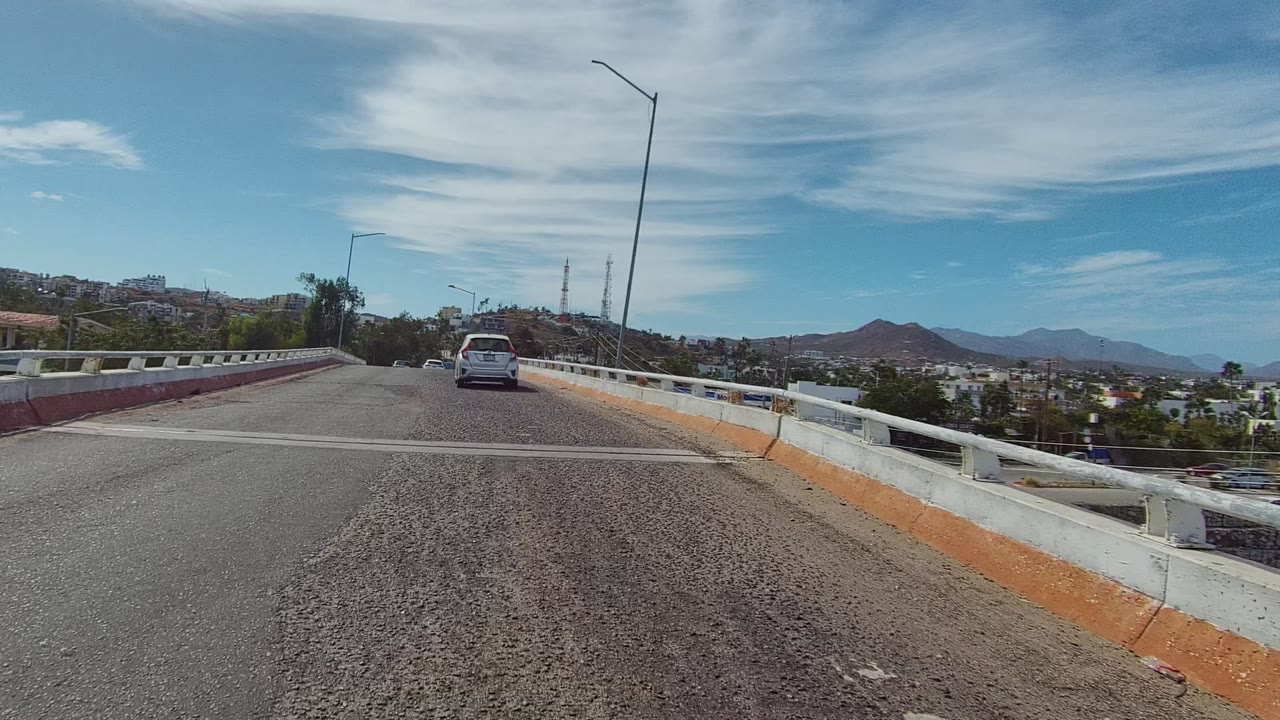 A car drives ahead on a bridge with guardrails and mountains beyond.