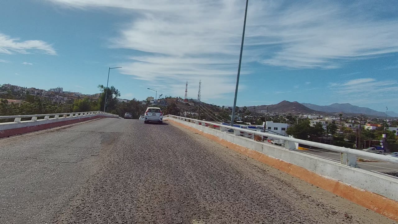 The vehicle is farther ahead on the Los Cabos bridge road, with rough asphalt filling the foreground, a tall streetlight at right, hills, low buildings, and no visible people.