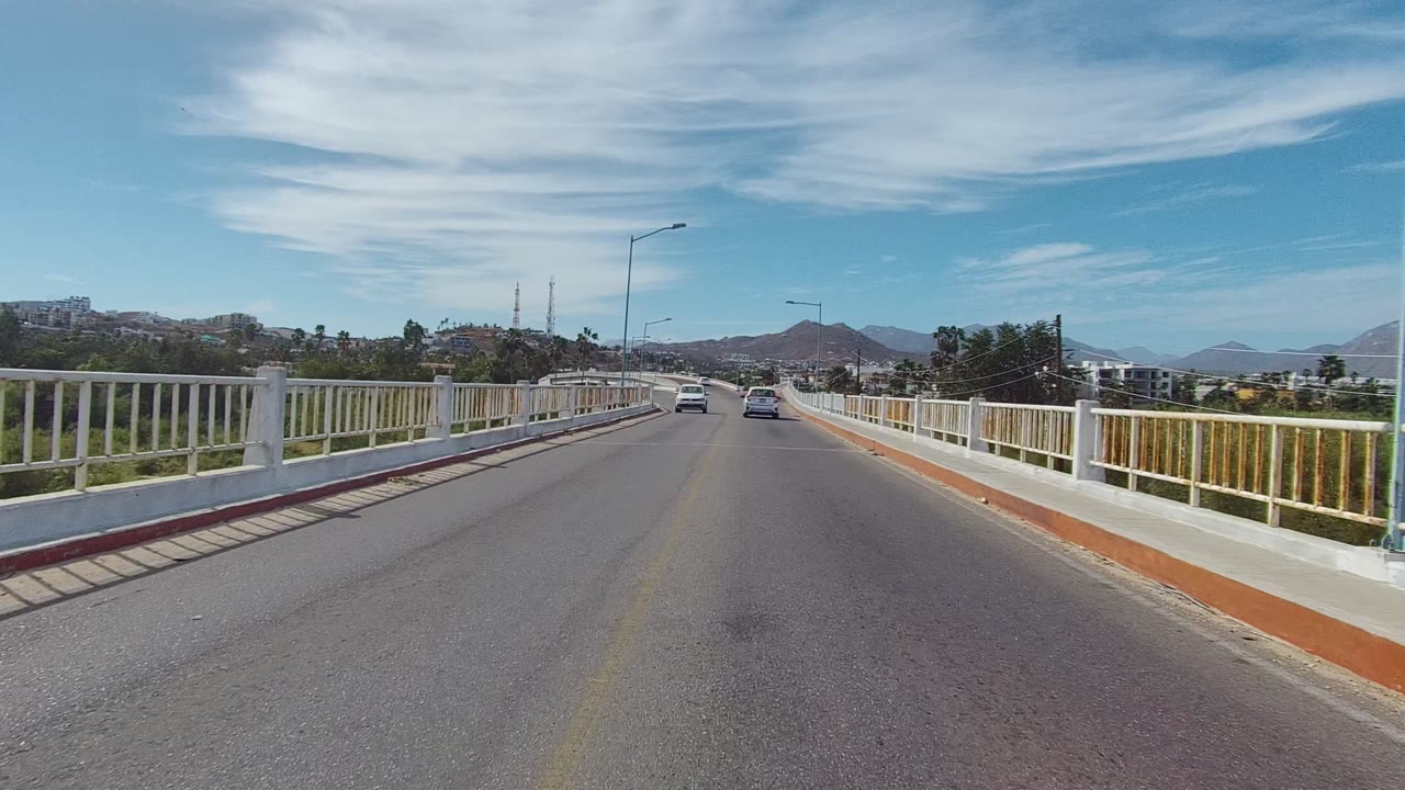 Forward road view on a bridge with white railings, cars ahead, mountains, and blue sky.