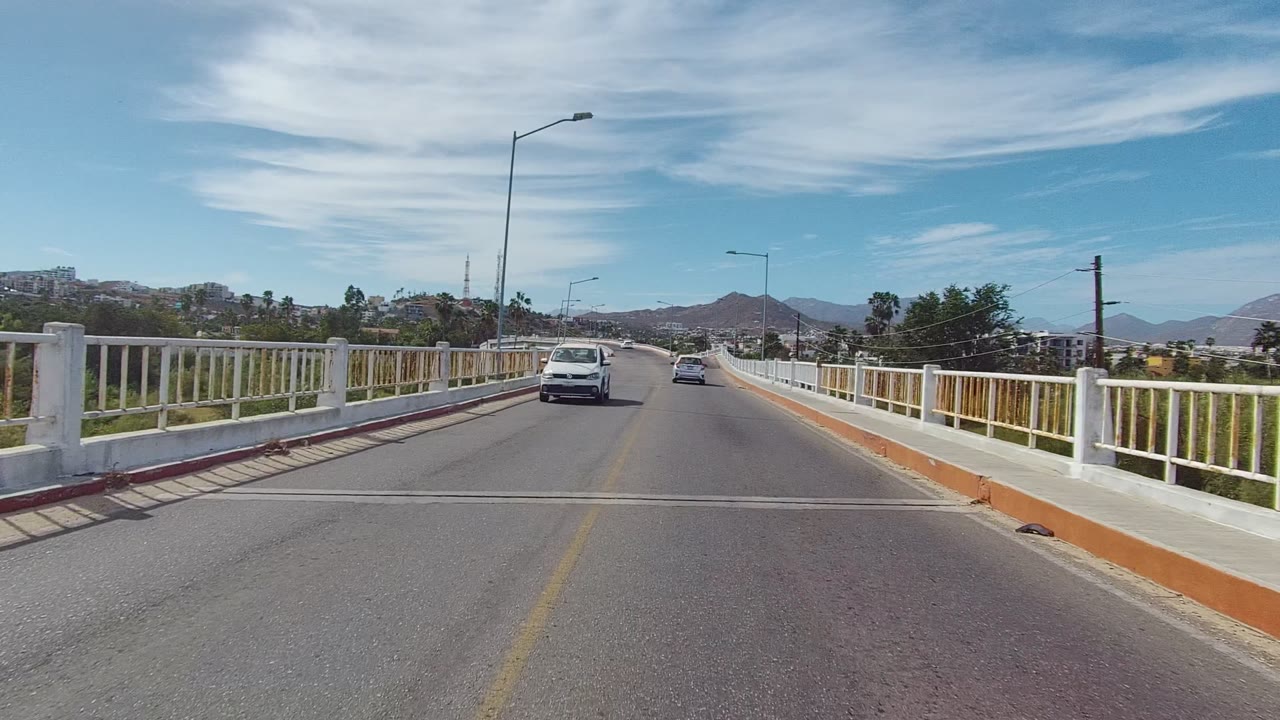 A later bridge-road frame in Los Cabos shows a white car nearer in the lane ahead, white railings and orange curbs along the sides, hills on the horizon, and no visible people.