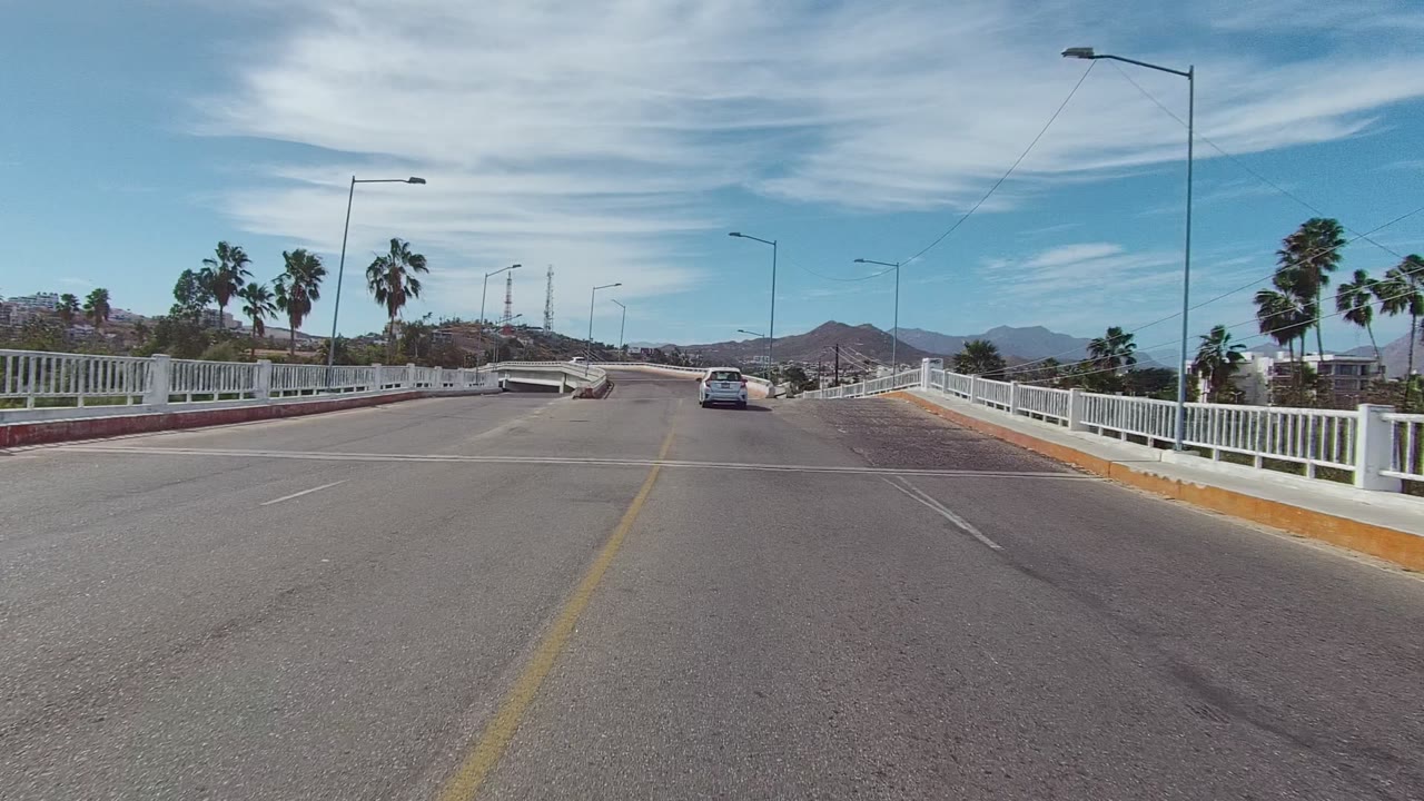 Vehicle-height view on a bridge road in East Cape Los Cabos, Baja California Sur, with white railings on both sides, cars ahead, palm trees, distant hills, and no visible people.