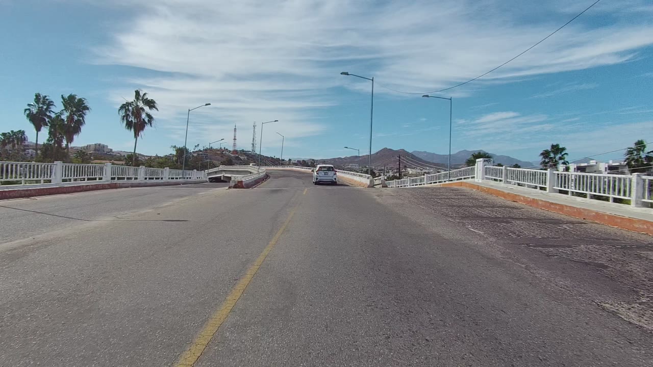 Roadway curves across a bridge with a car ahead, white rails, palms, and low mountains.