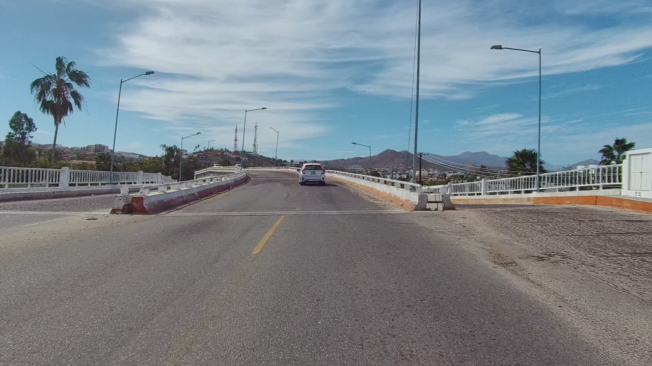 The East Cape Los Cabos bridge view continues with a wider roadside edge, white railings, cars ahead, tall streetlights, palm trees, hilly terrain, and no visible people.