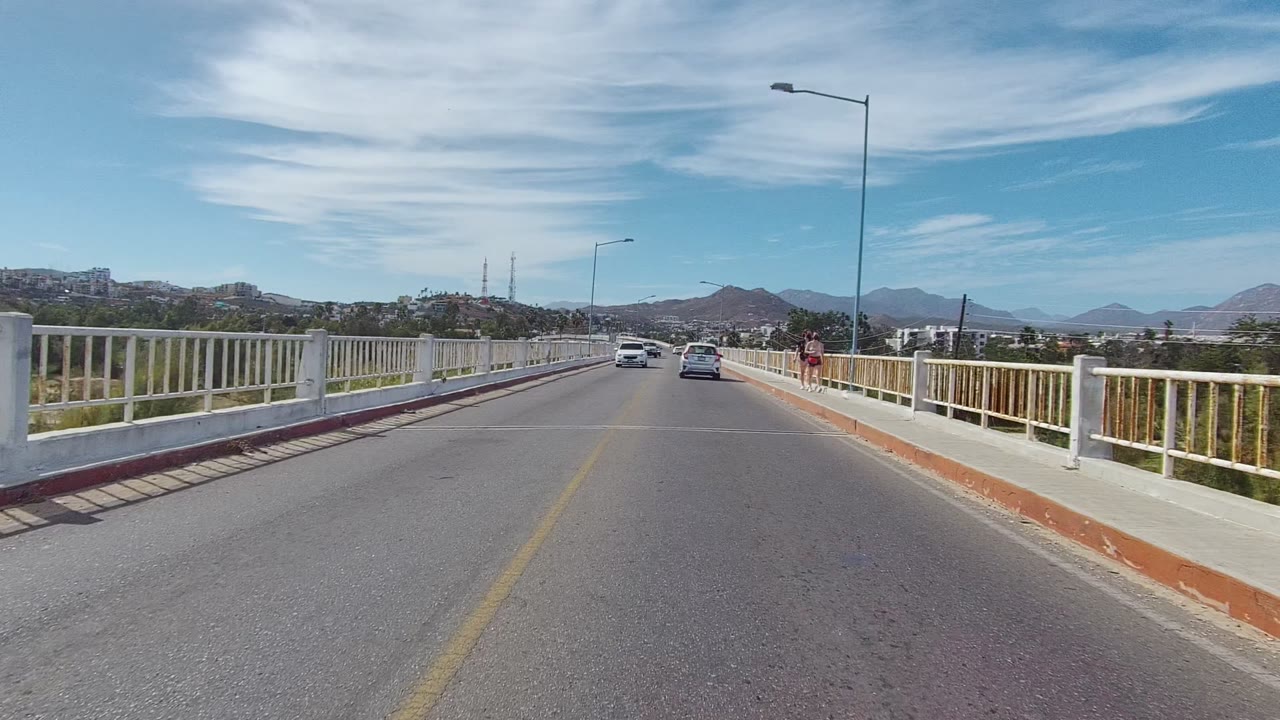 Road-level view on a bridge in Los Cabos, Baja California Sur, with cars ahead, white railings on both sides, distant mountains, and no visible people in this first frame.
