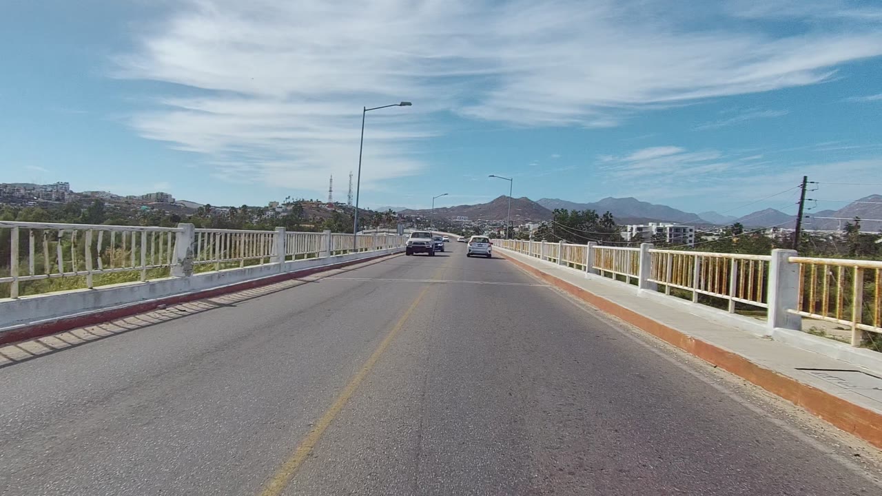 Forward view along the Los Cabos bridge road with cars farther ahead, white railings and red curbs framing the lane, mountains beyond, and no visible people in this frame.
