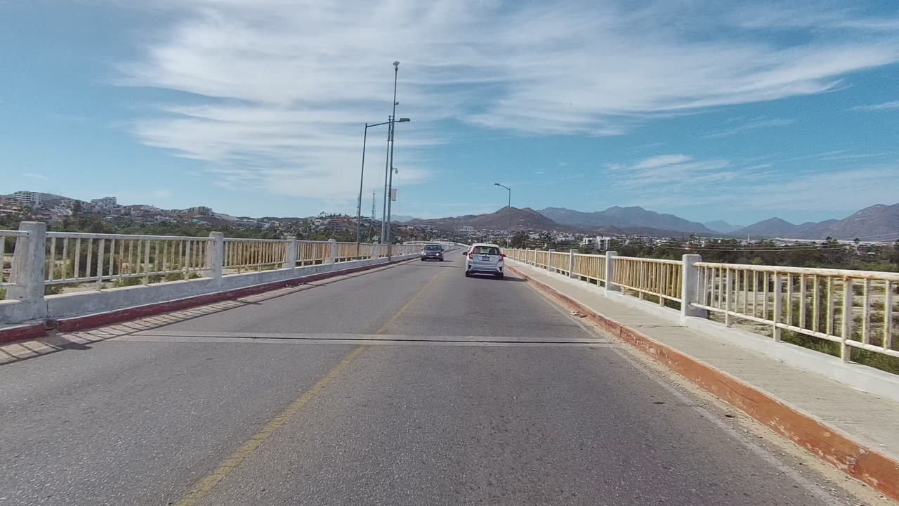 Start of the East Cape Los Cabos bridge view, with a white car ahead, pale railings on both sides, distant mountains, high clouds, and no visible people.