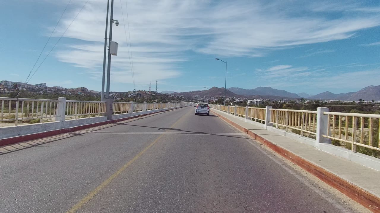 Mid-clip bridge view in East Cape Los Cabos, with a white car ahead in the lane, open railings along the road, mountain ridges across the horizon, and no visible people.