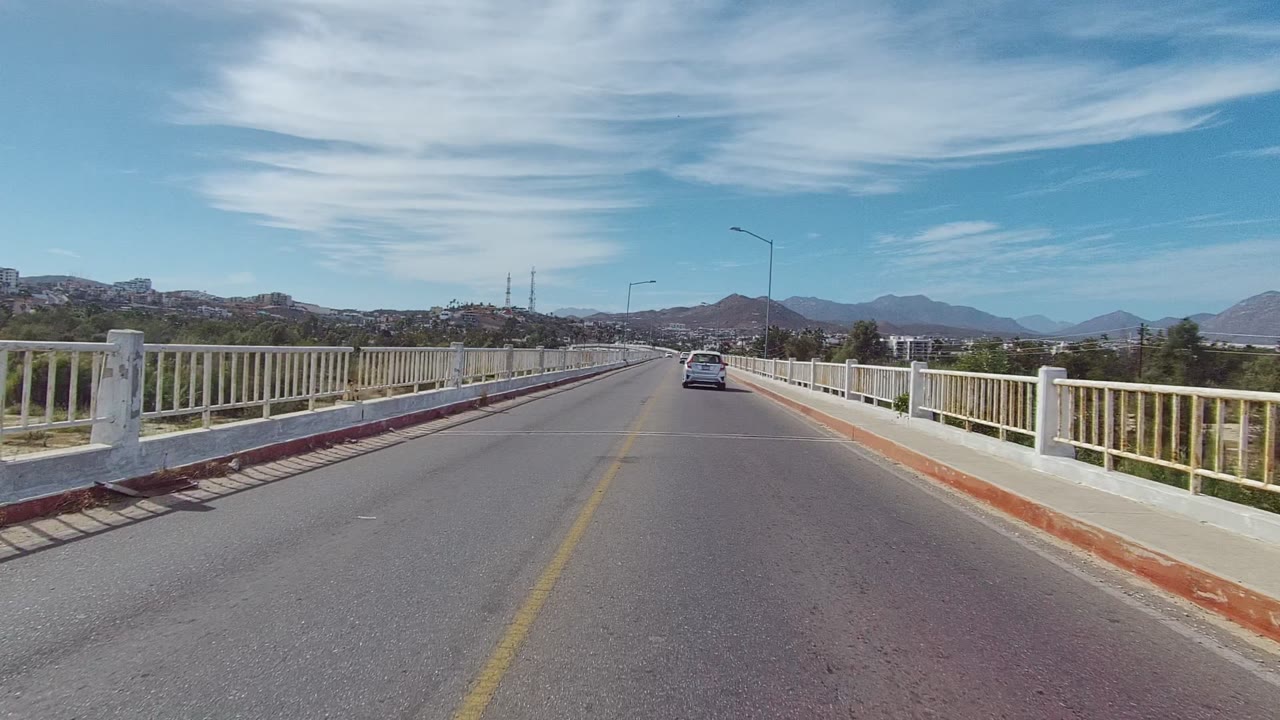 At 0:00, a car travels away on a bridge road in East Cape Los Cabos, with white railings, red curbs, a yellow center line, distant mountains, blue sky, and no visible people.