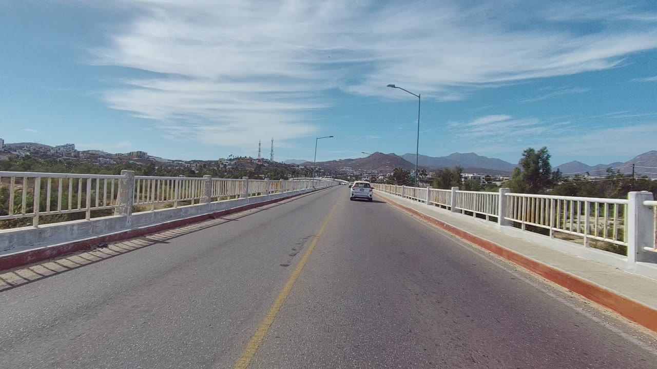 Bridge roadway with a car ahead, railings on both sides, and mountains beyond.