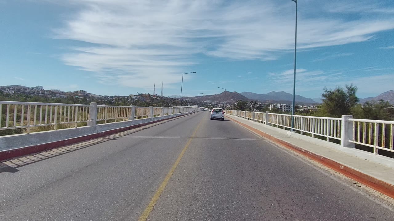 At 0:02, the East Cape Los Cabos road view holds the car ahead, a long right-side bridge railing, red curb edges, streetlights, mountains, and no visible people.