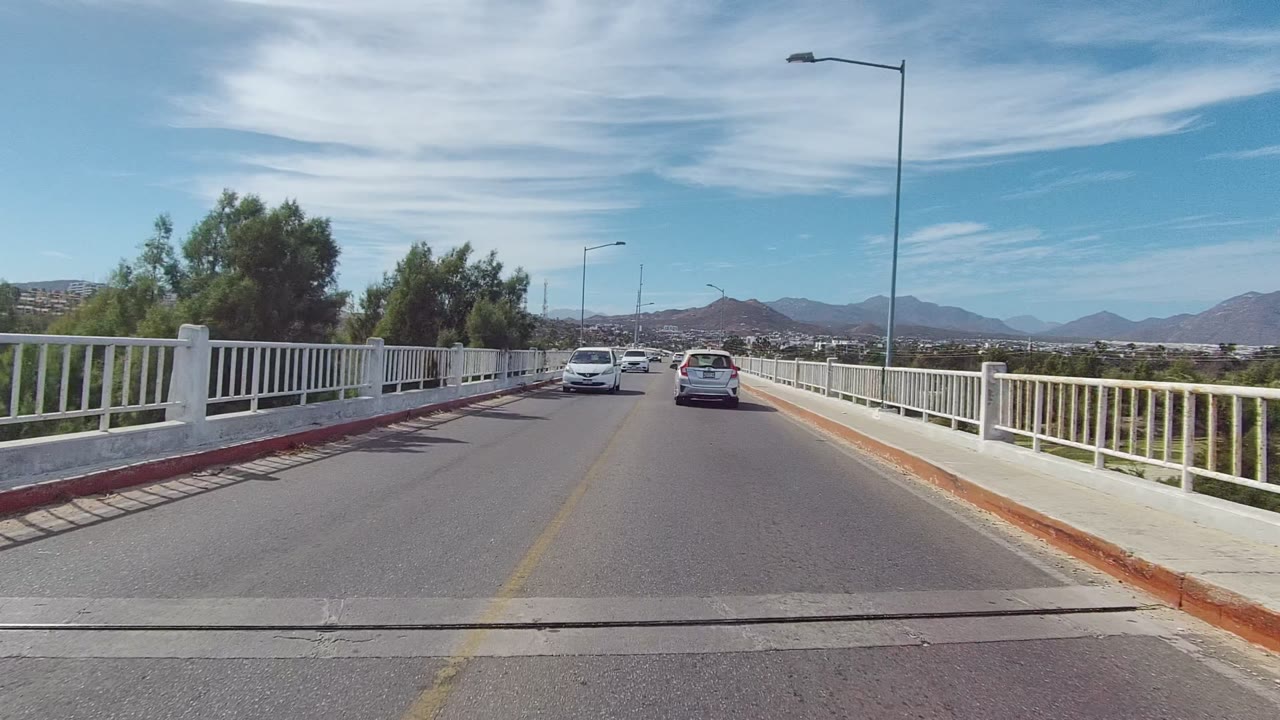Start of the East Cape Los Cabos clip, showing cars ahead on a two-lane bridge with white railings, streetlights, trees, distant mountains, blue sky, and no visible people.
