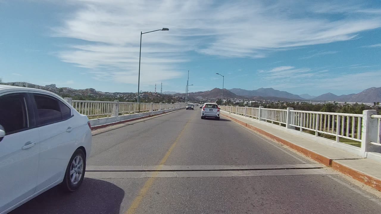 At 0:02 in the East Cape Los Cabos footage, a white car passes close on the right edge while another car continues ahead on the bridge, with railings, mountains, sky, and no visible people.