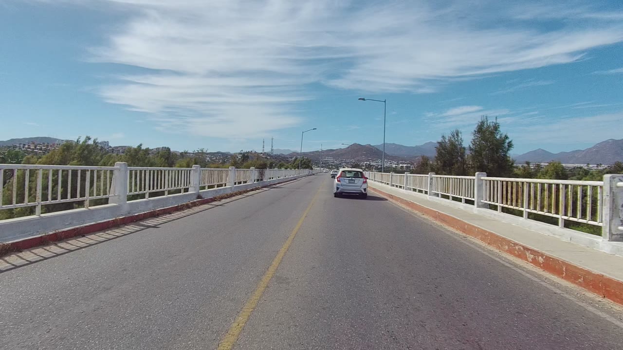A white car travels away from the camera on a bridge in East Cape Los Cabos, Mexico, with white railings, streetlights, distant mountains, blue sky, and no visible people.
