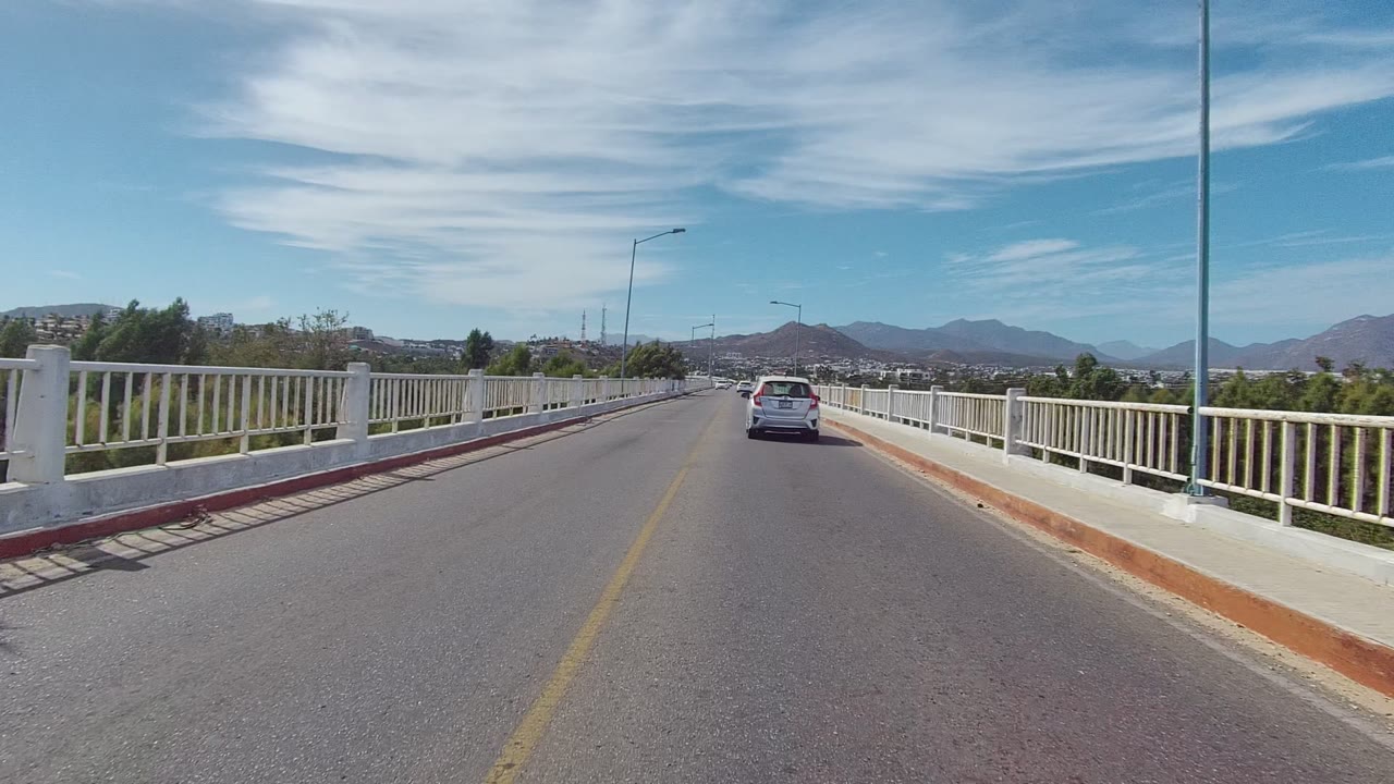 At 0:02, the white car is farther along the Los Cabos bridge, framed by white side railings, a reddish curb edge, streetlights, trees, mountains, and no visible people.