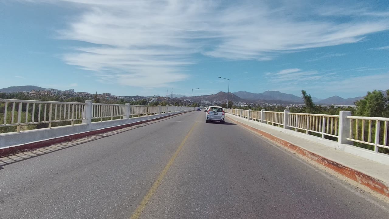 A white car drives ahead on a bridge road in East Cape Los Cabos, Baja California Sur, with white railings, streetlights, distant mountains, streaked clouds, and no visible people.