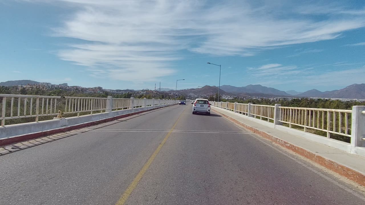 Bridge road with a white car ahead, railings, streetlights, and mountains in the distance.