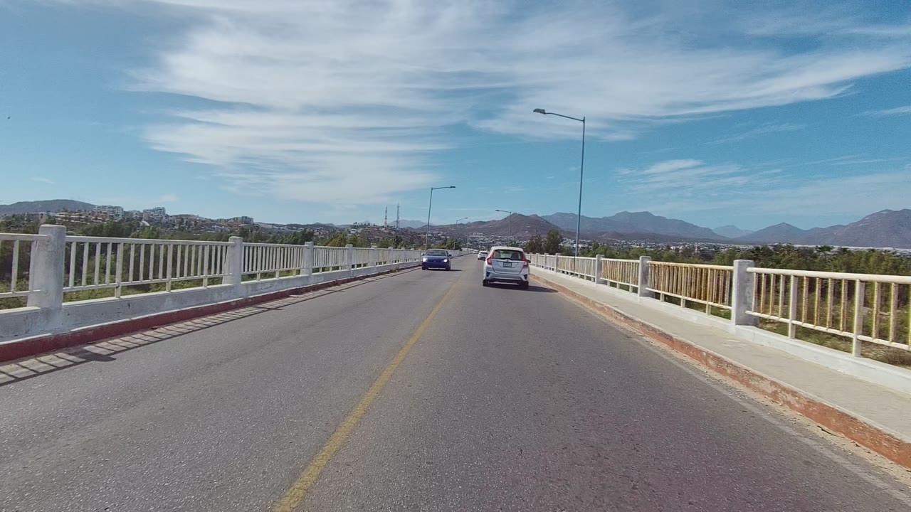 A white car remains ahead on the East Cape Los Cabos bridge road, with the right railing close to frame, mountains on the horizon, overhead streetlights, streaked clouds, and no visible people.