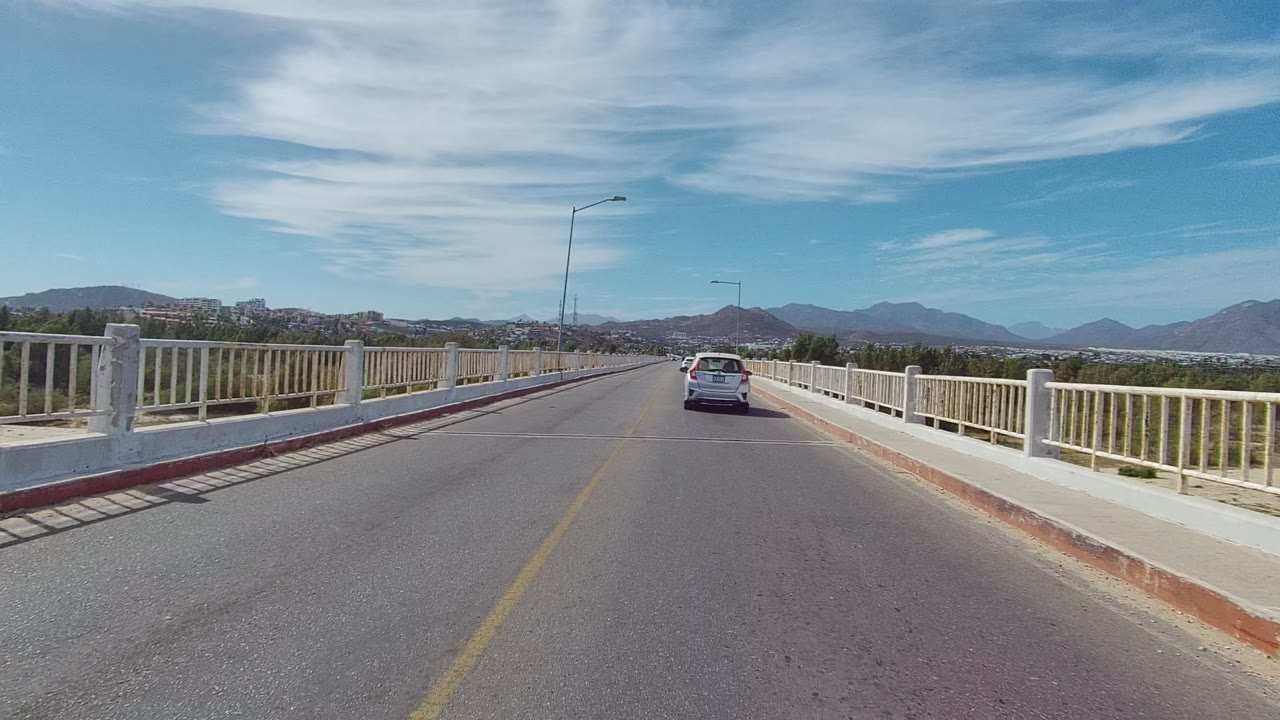 Opening view in East Cape Los Cabos, Mexico, with a car ahead on a bridge, white railings on both sides, distant mountains, scattered clouds, and no visible people.