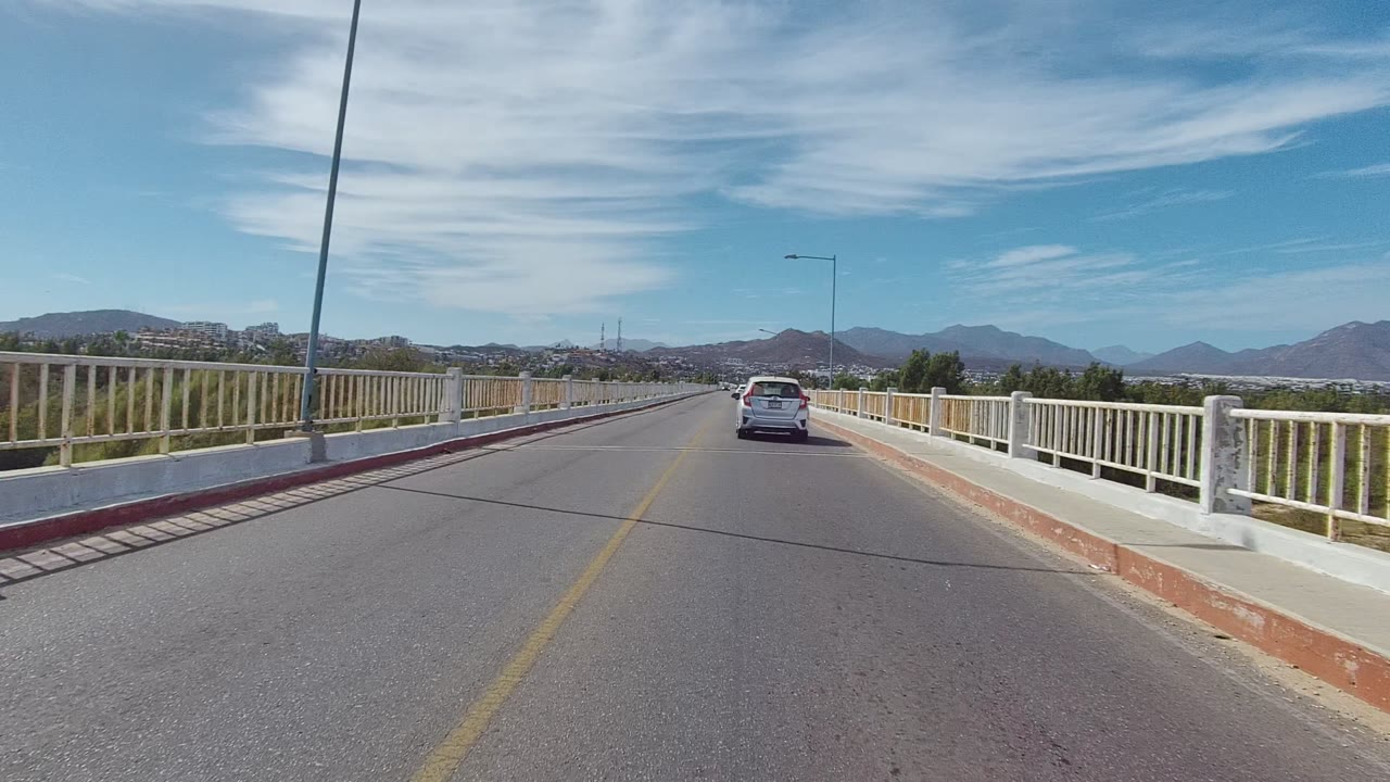 At 0:02, the East Cape Los Cabos roadway view shows the car farther along the bridge, with the right railing prominent, a tall streetlight overhead, mountains, and no visible people.