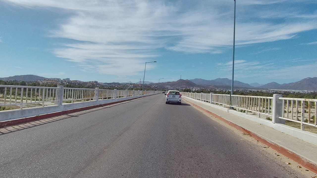 A white car drives away on a bridge road in East Cape Los Cabos, Mexico, with white railings, streetlights, distant mountains, blue sky, and no visible people.