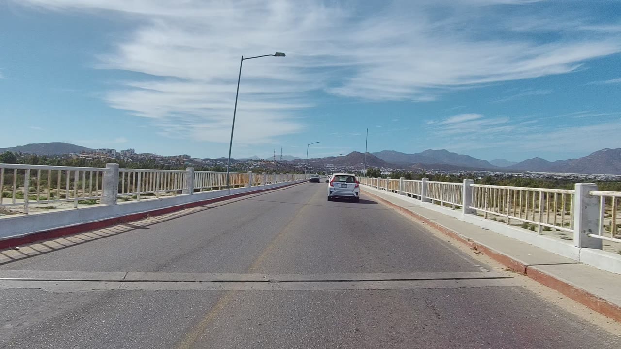 A white car remains ahead on the East Cape Los Cabos bridge road, framed by white side railings, red curb paint, road surface, distant hills, and no visible people.