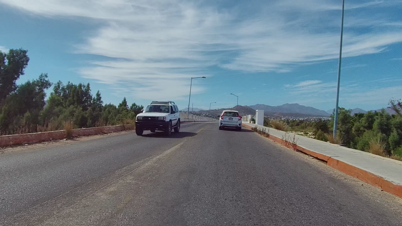 Opening frame in East Cape Los Cabos shows a white sedan ahead and a pickup passing on the left side of a bridge road, with railings, trees, mountains, and no visible people.