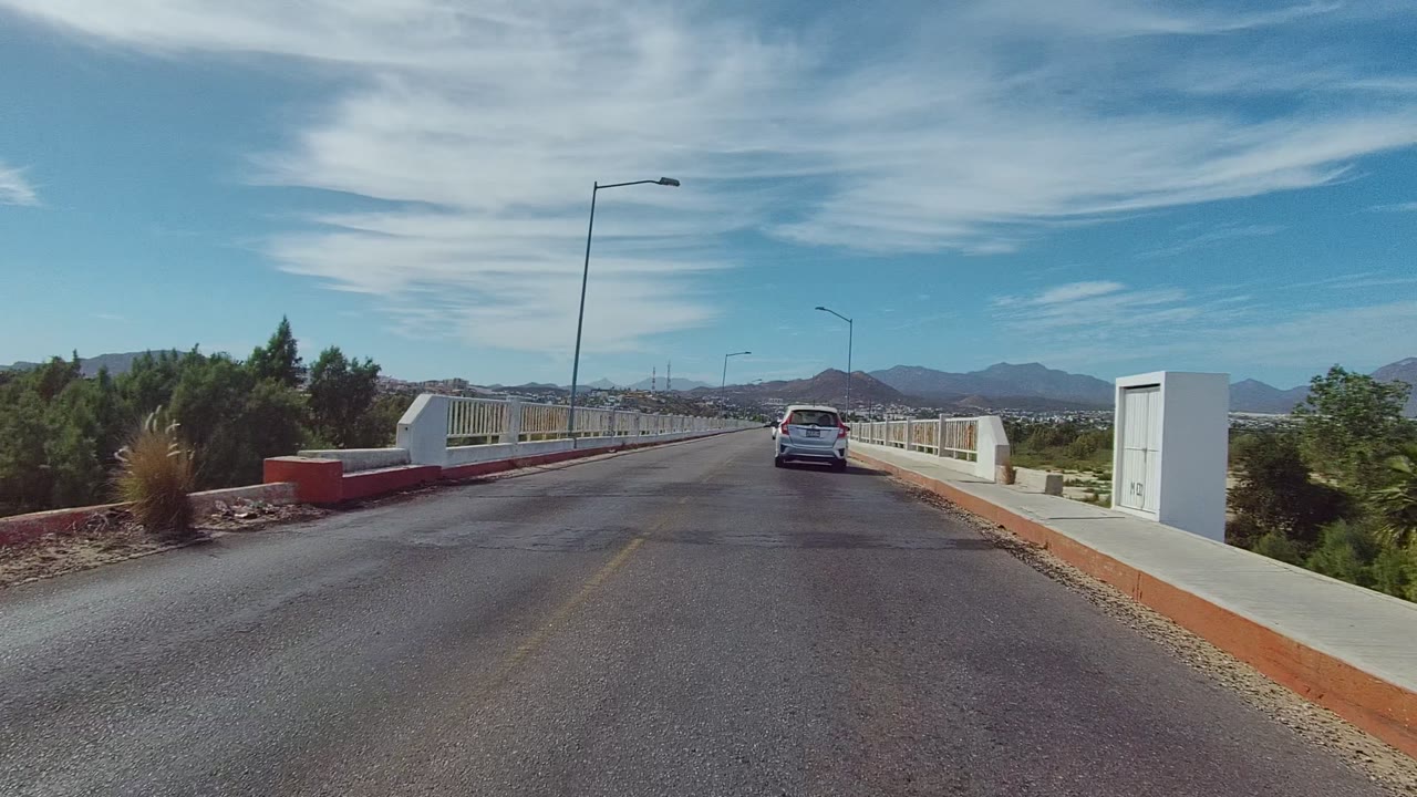 The road curves onto the bridge in East Cape Los Cabos as the white sedan moves farther ahead, with white railing posts, green shrubs, dry terrain, mountains, and no visible people.
