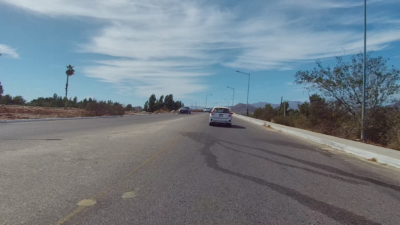 Vehicle-perspective view of a paved road in East Cape Los Cabos, Mexico, with a white car ahead, dry roadside vegetation, palm trees, distant hills, thin clouds, and no visible people.