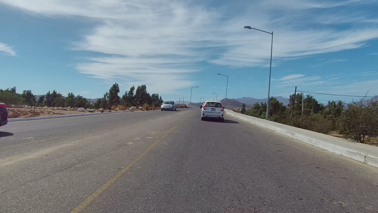 East Cape Los Cabos road view with a white car ahead, a dark vehicle at the left edge, dry shrubs, palms, streetlights, mountains on the horizon, and no visible people.