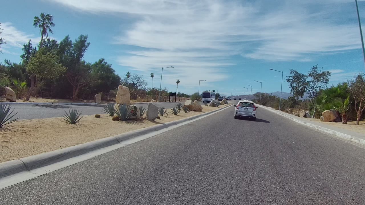 At 0:00 in East Cape Los Cabos, a white car drives ahead on a paved road bordered by sandy landscaping, boulders, shrubs, streetlights, distant palms, and no visible people.