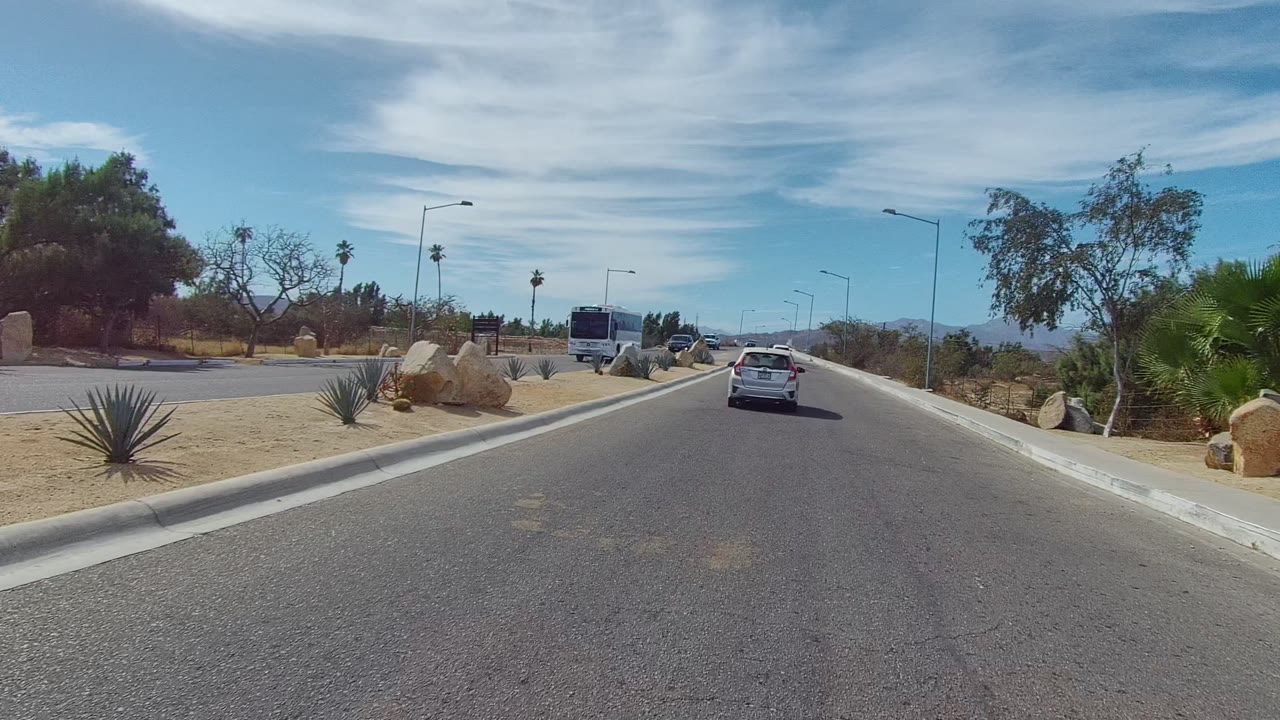 White car on a paved road near roadside rocks, shrubs, and a bus in the distance.
