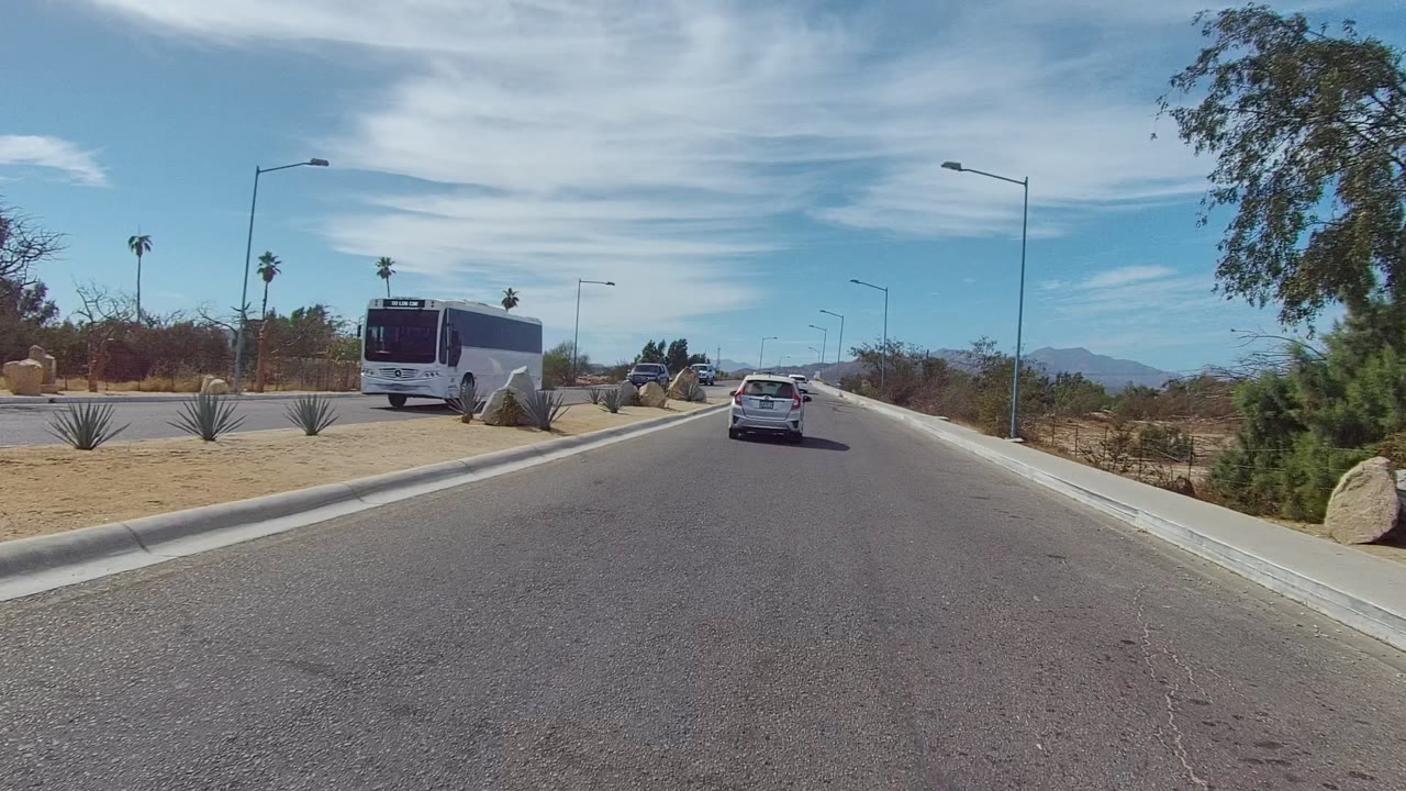 At 0:02 in East Cape Los Cabos, a bus passes near the left edge as a white car travels ahead on the paved road beside boulders, dry plants, streetlights, and no visible people.