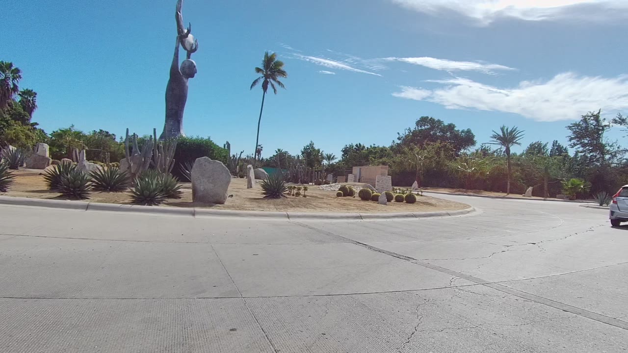 Opening frame of an East Cape Los Cabos roundabout with a tall statue, rocks, cacti, palms, dry planting beds, broad pavement, and no visible people.