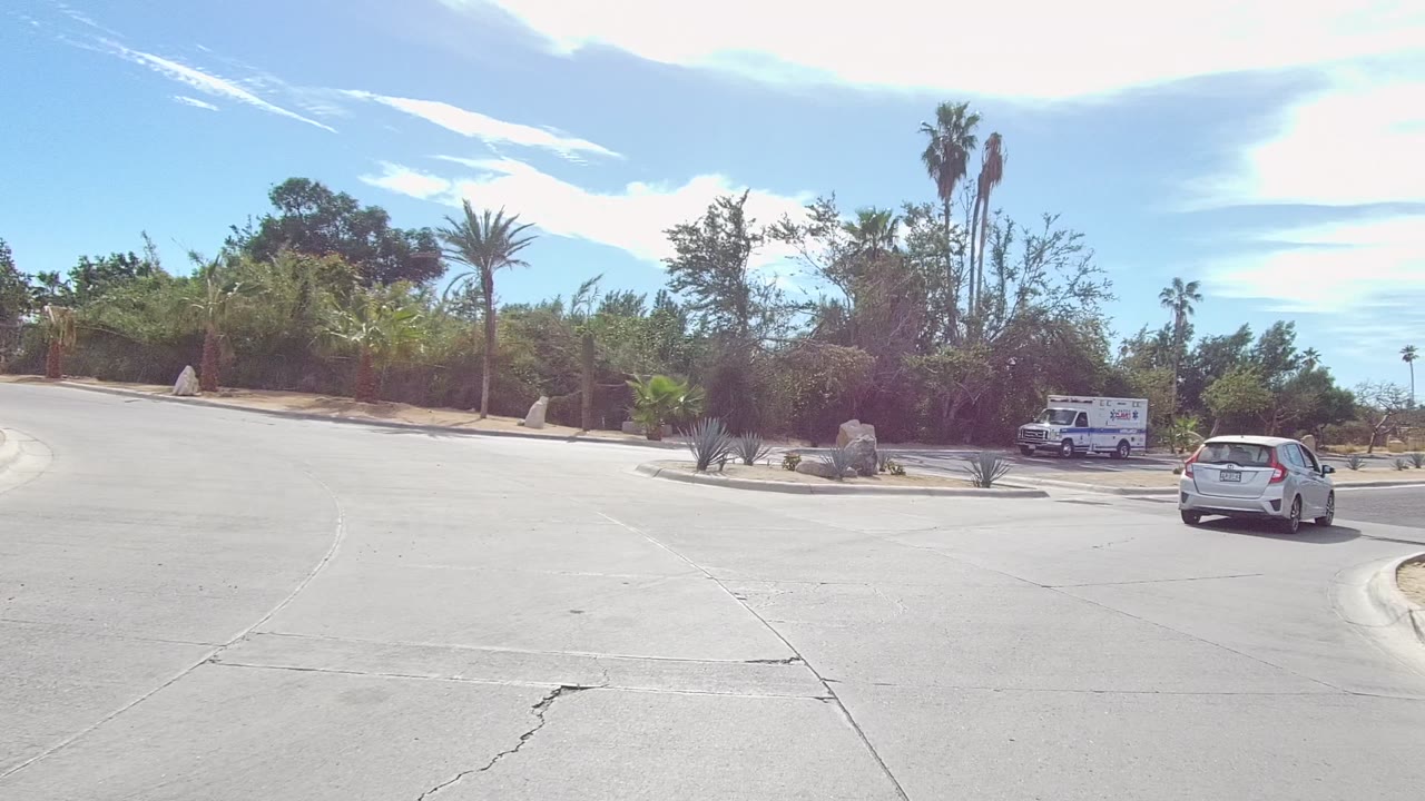 The East Cape Los Cabos road curves around the traffic island as a white car and a marked ambulance sit near rocks, palms, shrubs, bright sky, and no visible people.