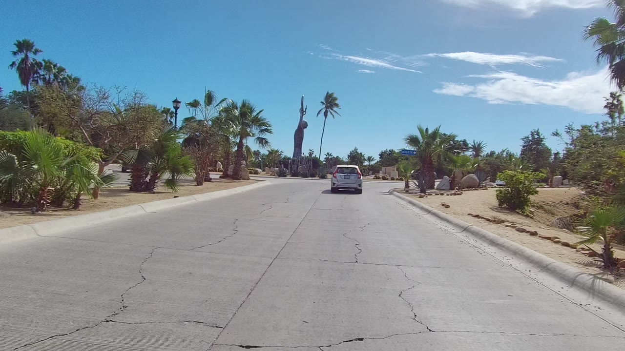 A Los Cabos roadway curves near street signs, palms, desert plantings, a tall statue in the distance, and a white car at right, with no visible people.
