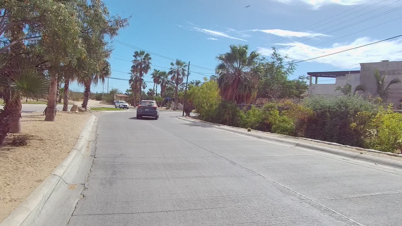 East Cape Los Cabos residential road with a dark car ahead, sandy shoulders, palms, overhead wires, and no visible people.