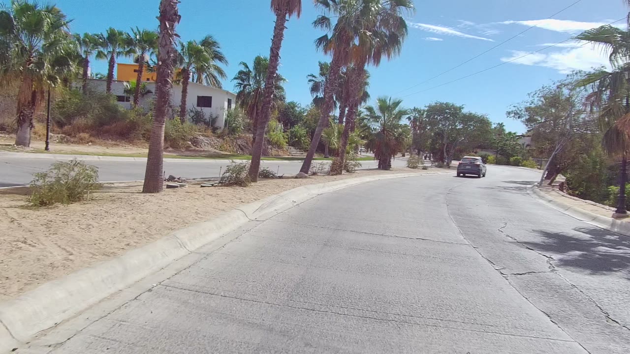 Opening frame of an East Cape Los Cabos road with sandy shoulders, palms on both sides, a dark car ahead, low buildings in the distance, and no visible people.