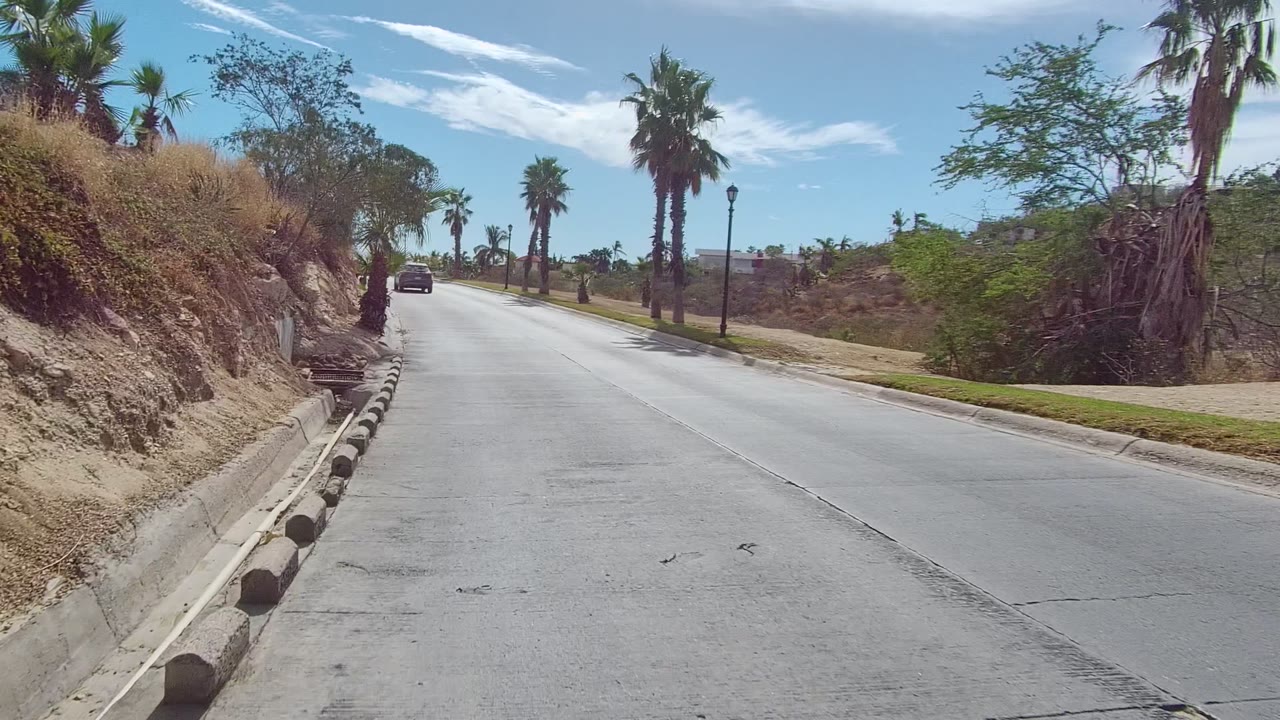 At 0:00 in East Cape Los Cabos, a concrete road rises between a dry cut hillside and palms, with a car ahead and no visible people.