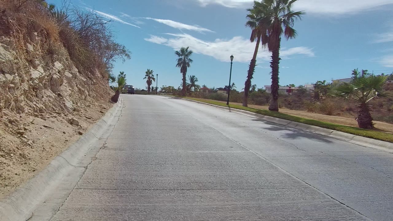 At 0:02 in East Cape Los Cabos, a curving concrete road passes a rocky dry hillside on the left with palms, lamp posts, and no visible people.